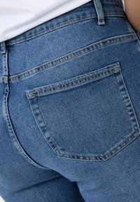 Jean en denim bleu taille haute avec une seule poche arrière, des coutures visibles et une texture lisse. Le tissu semble légèrement extensible.