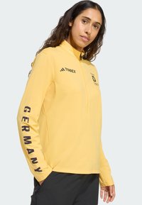 Geel atletisch shirt met lange mouwen en een halve rits, met "GERMANY" in het zwart op de mouw; gemaakt van flexibel, ademend materiaal.