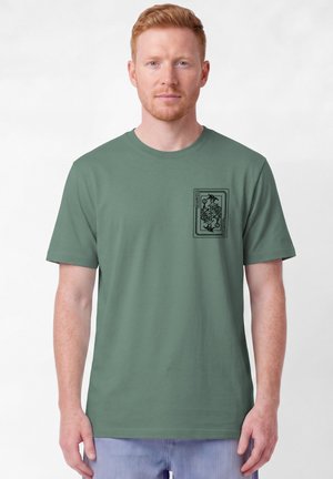 Mann mit kurzen roten Haaren, der ein grünes T-Shirt mit einer schwarzen Joker-Spielkarte-Grafik auf der oberen linken Brust trägt.