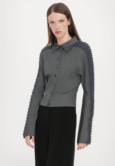 Grå cropped skjorte med knapfront, spids krave og tekstureret stof langs ærmerne. Lange ærmer har et zigzag-mønster.