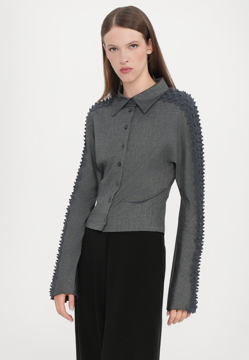 Grå kortärmad skjorta med knappstängning, spetsad krage och strukturerad tyg längs ärmarna. Långa ärmar har ett zickzackmönster.