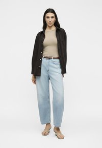 Γυναίκα στέκεται φορώντας μπεζ ριγέ τοπ, ανοιχτό μαύρο πουκάμισο, ανοιχτό μπλε τζιν, καφέ ζώνη και μπεζ σανδάλια σε λευκό φόντο.