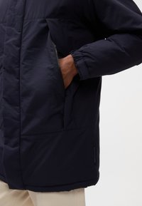 Giacca a giacca blu navy con tasca con zip, polsini a coste e texture liscia. Presenta una vestibilità leggermente oversize e materiale isolante.
