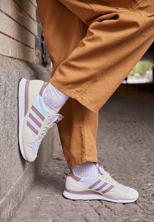 Osoba nosząca białe i różowe buty sportowe Adidas oraz białe skarpetki, z jedną stopą opartą o kamienny mur na brukowanej ulicy.