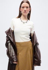 Γυναίκα που φοράει λευκή μπλούζα με κοντά μανίκια, κίτρινη φούστα με καρό σχέδιο, ασημένιο κολιέ αλυσίδα, κρεμαστά σκουλαρίκια και σκούρο καφέ δερμάτινο σακάκι περασμένο στους βραχίονες.