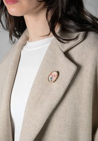 Bežna volnena bunda z izrezanim ovratnikom, ki na reverju nosi cvetlično broško v zlatem okviru. Pod njo je bela pulover, ki dodaja teksturo.