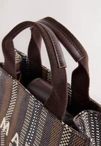 Sac fourre-tout avec un motif tissé à rayures marron et crème, avec des accents en cuir marron et des poignées en tissu robustes. L'ouverture montre l'intérieur.