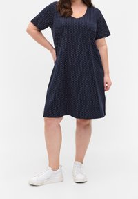 Kobieta ubrana w granatową sukienkę do kolan w białe kropki i białe trampki, stojąca, opierając jedną rękę na biodrze.
