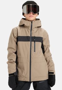 Osoba v béžovej a čiernej lyžiarskej bunde, čiernych rukaviciach, čiernej čiapke a reflexných lyžiarskych okuliaroch na neutrálnom pozadí.