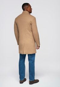 Beige Übermantel mit hohem Kragen, einem Schlitzausschnitt hinten, getragen über blauen Jeans und braunen Stiefeln. Glatte Textur, taillierte Passform, dezente Knöpfe.