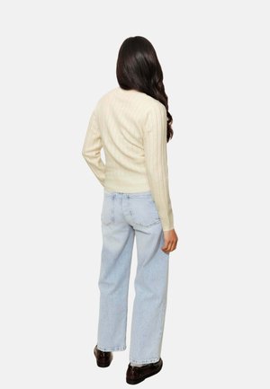 Pull en maille côtelée crème à manches longues, associé à un jean large bleu clair et des chaussures foncées, vu de dos.