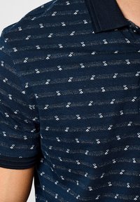 Polo shirt blu navy con motivo a maglia testurizzata e disegni geometrici bianchi. Il colletto è di un blu navy solido e la maglietta ha maniche corte.
