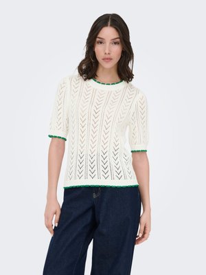 Femme portant un pull en maille blanc à manches courtes avec une bordure verte et un jean large foncé, debout sur un fond clair uni.