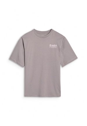 Młoda osoba nosząca szary oversize t-shirt, czarne spodnie, czarne trampki i szarą czapkę noszoną do tyłu, stojąca z jedną ręką przy głowie.