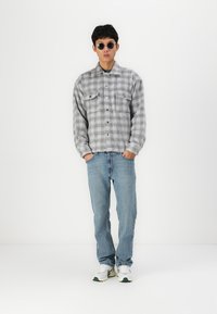 Grijs en zwart geruite blouse met twee borstzakken, blauwe jeans en witte sneakers met groene accenten. Draagt ronde zonnebril.