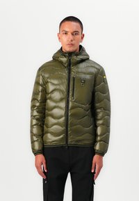 Veste en duvet isolante vert olive avec fermeture éclair, capuche et design matelassé. Elle comporte une poche poitrine et des poignets côtelés.