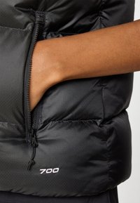 Svart pufferväst med texturerad yta, utrustad med en ficka med dragkedja och broderad "700"-detalj nära nederkanten.