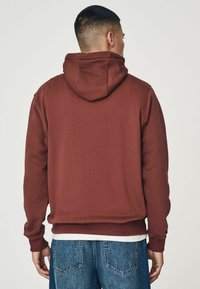 Sudadera burdeos con un acabado texturizado, que presenta una capucha con cordón y puños acanalados. Combinada con unos vaqueros de mezclilla azul.