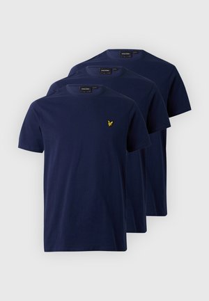 Man staat, draagt een donkerblauw t-shirt, lichtblauwe jeans, rode schoenen, zonnebril en een gouden ketting op een effen lichte achtergrond.