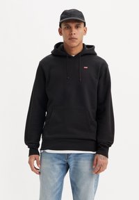 Zwarte hoodie van katoen, met een voorzak, trekkoordkap en klein rood logo. Draagt over lichtblauwe jeans.