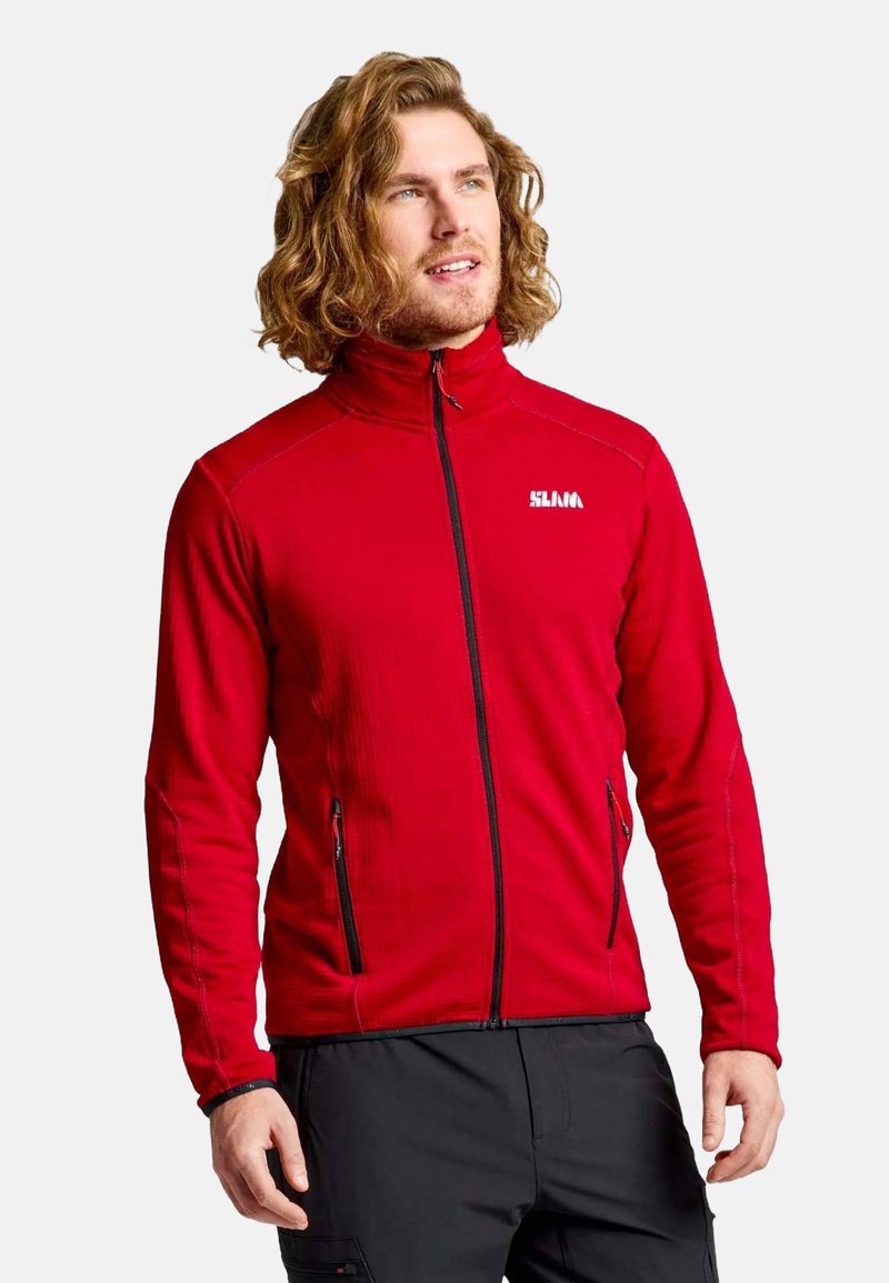 Homme aux cheveux bouclés portant une veste zippée rouge et un pantalon noir, se tenant contre un fond clair, regardant légèrement en haut et à droite.