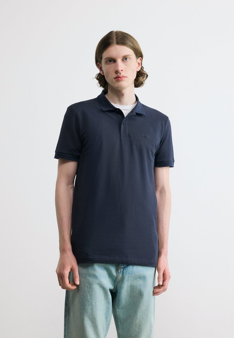 Νεαρός με μεσαίου μήκους καστανά μαλλιά, φοράει μπλε σκούρο πόλο πουκάμισο πάνω από λευκό κοντομάνικο και ανοιχτόχρωμα τζιν, στέκεται μπροστά σε ένα απλό φόντο.