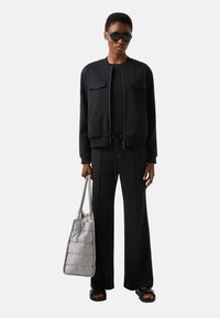 Czarna dzianinowa kurtka typu bomber z dwoma przednimi kieszeniami, zestawiona z czarnymi spodniami o szerokich nogawkach. Dodatki to szara pikowana torba i okrągłe okulary przeciwsłoneczne.