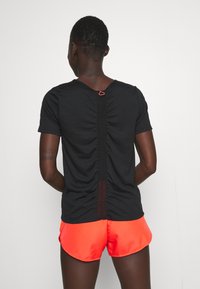 Person med kort hår sedd bakifrån, iklädd en svart t-shirt med rynkad rygg och klara orangea sportshorts mot en enfärgad bakgrund.