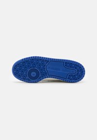 Tălpile de sneakers albi cu un fund negru din cauciuc texturat, având modele circulare și un logo embossat. Formă plată și rotundă cu un design de aderență.
