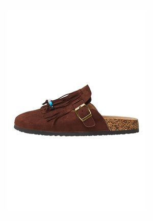 Chaussure brune en daim à enfiler avec des franges décoratives et de petites perles. Semelle intermédiaire en liège et semelle en caoutchouc visible, avec un détail de boucle sur le côté.