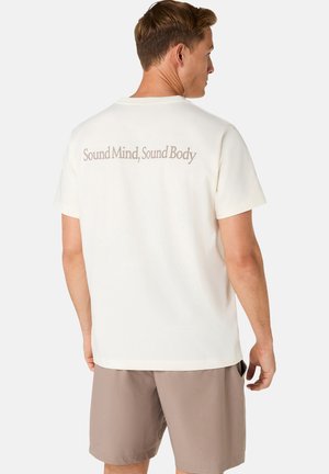 Mężczyzna w białej koszulce z napisem „Sound Mind, Sound Body” na plecach i beżowych szortach, stojący na jednolitym tle.