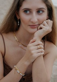 Bijuterii din aur cu lanțuri suprapuse, perle și inele texturate. Model cu piele deschisă la culoare și păr lung sprijină bărbia în mână, afișând accesoriile.