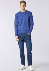 Maglione blu a maglia con scollo rotondo, maniche lunghe e logo sul petto, abbinato a jeans in denim scuro e sneakers bianche.