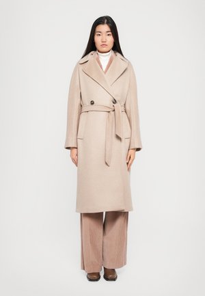 Cappotto in misto lana beige con design doppiopetto, ampio colletto, cintura in vita e tasche laterali, indossato sopra pantaloni in corduroy a gamba larga.