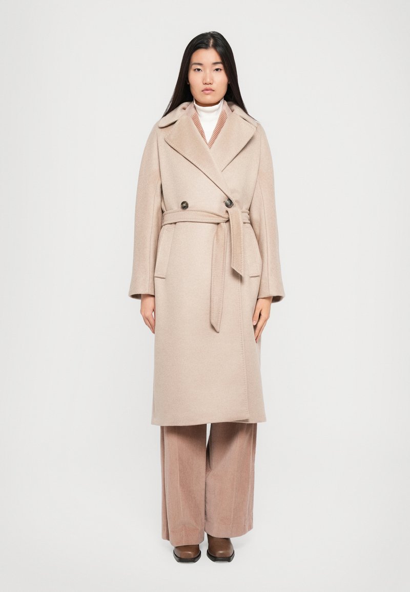 Beige wool blend coat with a double-breasted design, large collar, waist tie, and side pockets, worn over wide-legged corduroy pants.