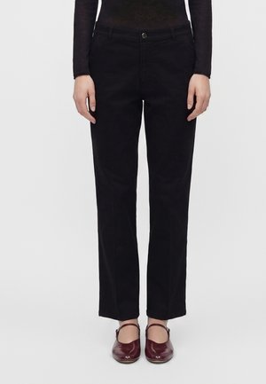 Persona che indossa pantaloni neri a gamba dritta, top nero a maniche lunghe e scarpe stile Mary Jane rosso scuro, in piedi davanti a uno sfondo semplice.