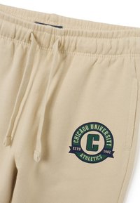 Beige joggingbroek met elastische tailleband en trekkoorden, voorzien van een groen en marineblauw logo "Chicago University Athletics" op de bovenbenen.