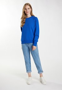 Blauwe crewneck sweatshirt van zacht materiaal, met ribgebreide manchetten en zoom. Gecombineerd met opgerolde lichtblauwe jeans en witte sneakers.