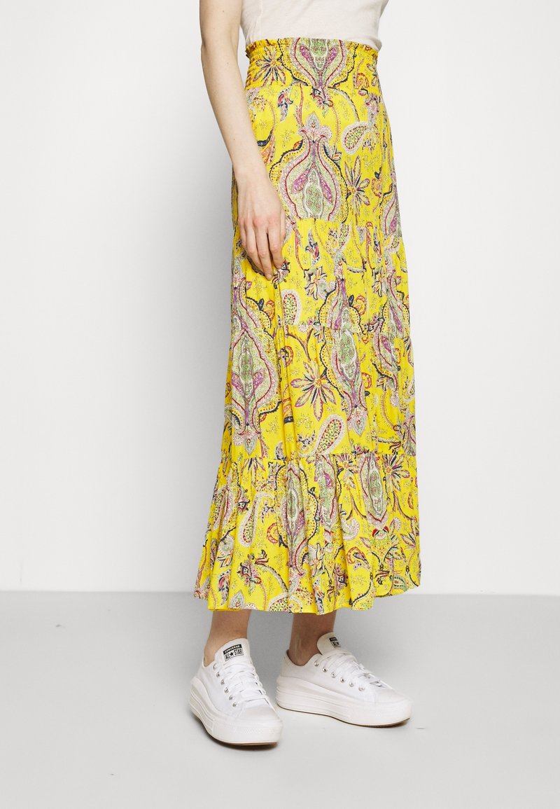 Falda larga amarilla con patrón paisley, cinturilla fruncida y dobladillo con volantes. Combinada con zapatillas blancas. La tela parece ligera y fluida.