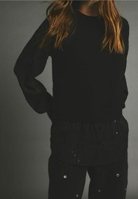 Pull-over noir à motif texturé, porté sur une chemise noire texturée à boutons ornée de sequins, assortie à un pantalon coordonné.