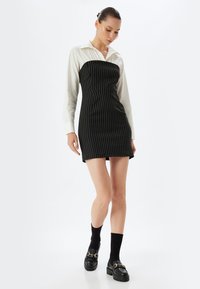 Schwarzes gestreiftes Kleid mit einem weißen Kragenhemd als Überwurf, das einen taillierten Oberkörper und einen kurzen Saum hat. Getragen mit schwarzen Plateau-Schuhen und Socken.