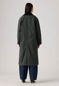 Lange, donkergroene trenchcoat met een zwarte kraag, knoopmanchetten en een split aan de achterkant. Draagt over brede blauwe jeans en zwarte hakken.