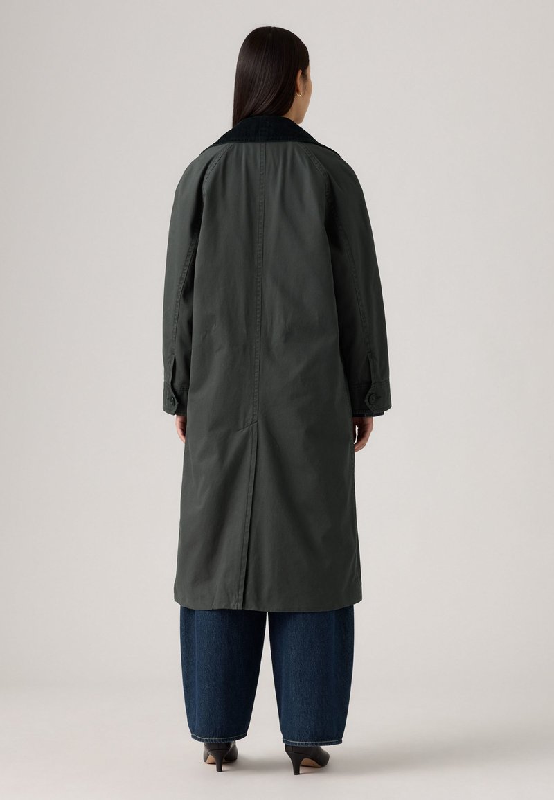 Long manteau vert foncé, avec un col noir, des poignets boutonnés et une fente à l'arrière. Porté sur un jean large bleu et des escarpins noirs.