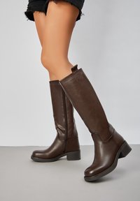 Bottes en cuir marron montantes avec une texture lisse, bout carré et petit talon bloc. Elles sont dotées d'une fermeture éclair latérale et d'une languette à l'arrière.
