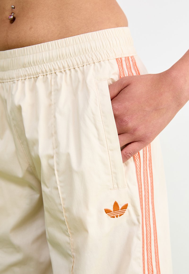 Jasno beżowe szorty z elastycznym pasem, ozdobione pomarańczowymi trzema paskami oraz haftowanym logo na bocznej kieszeni.