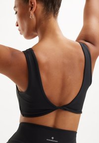 Crop top atletico nero con spalline larghe e scollatura profonda sulla schiena, realizzato in tessuto liscio con un dettaglio annodato sul retro.