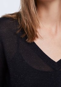 Vrouw met schouderlang lichtbruin haar die een zwarte V-hals trui draagt met subtiele glitterdraadjes.