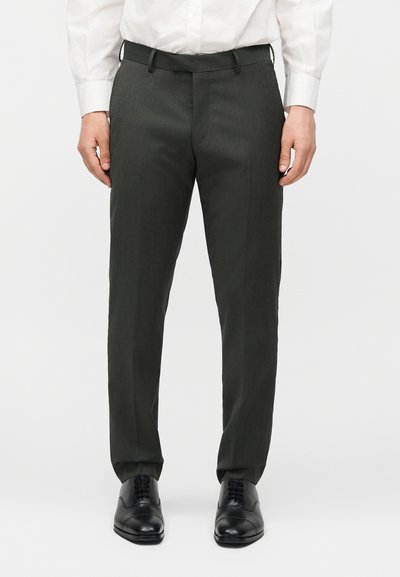 Άνδρας που φορά σκούρο γκρι επίσημο παντελόνι, μαύρα δερμάτινα παπούτσια και λευκό πουκάμισο μπουζωμένο μέσα στο παντελόνι, στέκεται σε απλό φόντο.