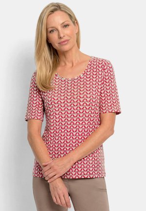 Femme blonde portant une chemise à manches courtes à motifs rouges, blancs et roses et un pantalon beige, debout les bras croisés.