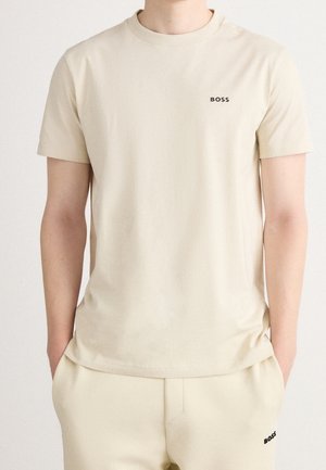 Beiges T-Shirt mit kurzen Ärmeln und passenden Hosen, beide mit kleinen schwarzen "BOSS"-Logos, Hände in den Taschen, neutraler Hintergrund.